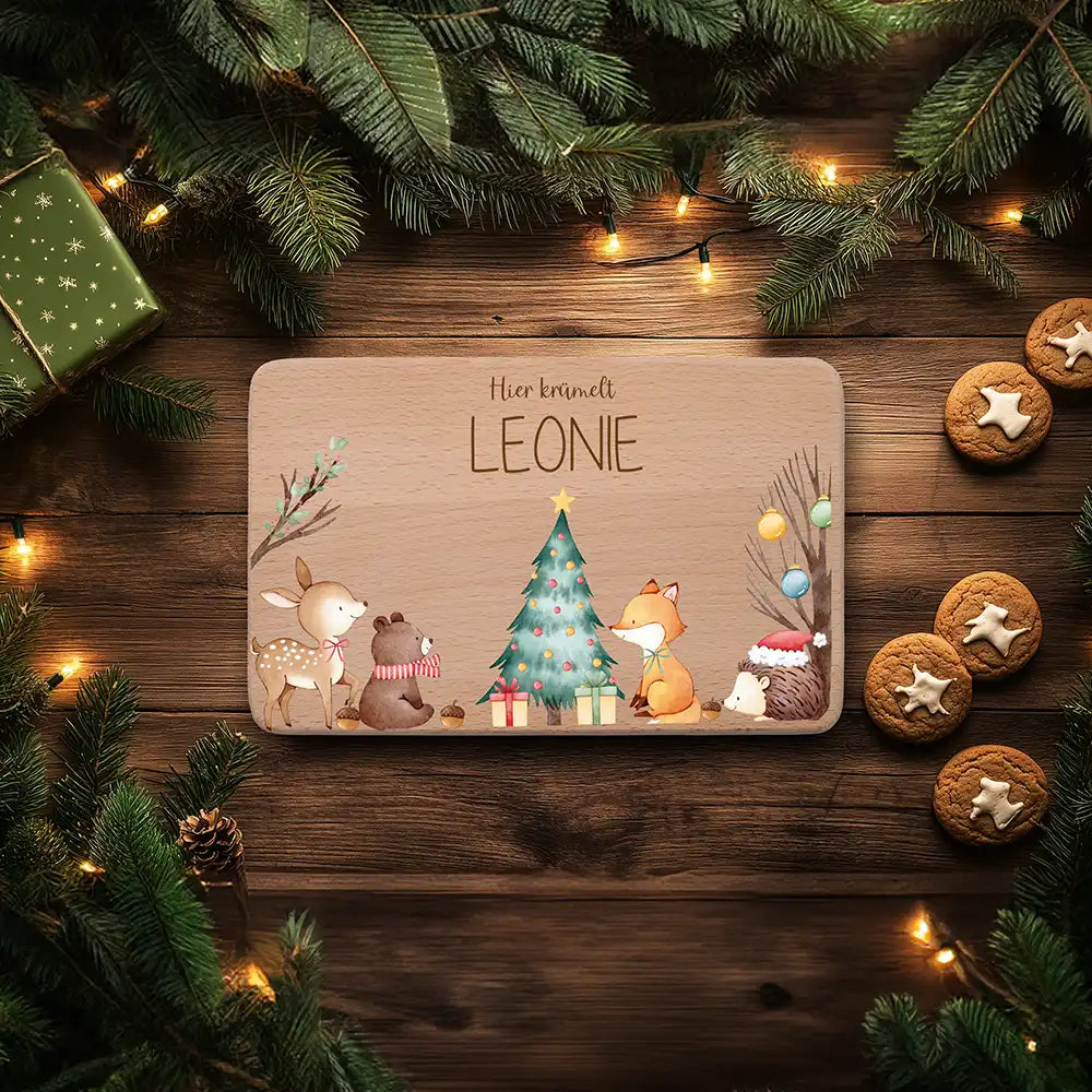 FrĂĽhstĂĽcksbrettchen mit dem Namen Leonie und Waldtieren um einen Weihnachtsbaum, daneben Tannenzweige und Lebkuchen