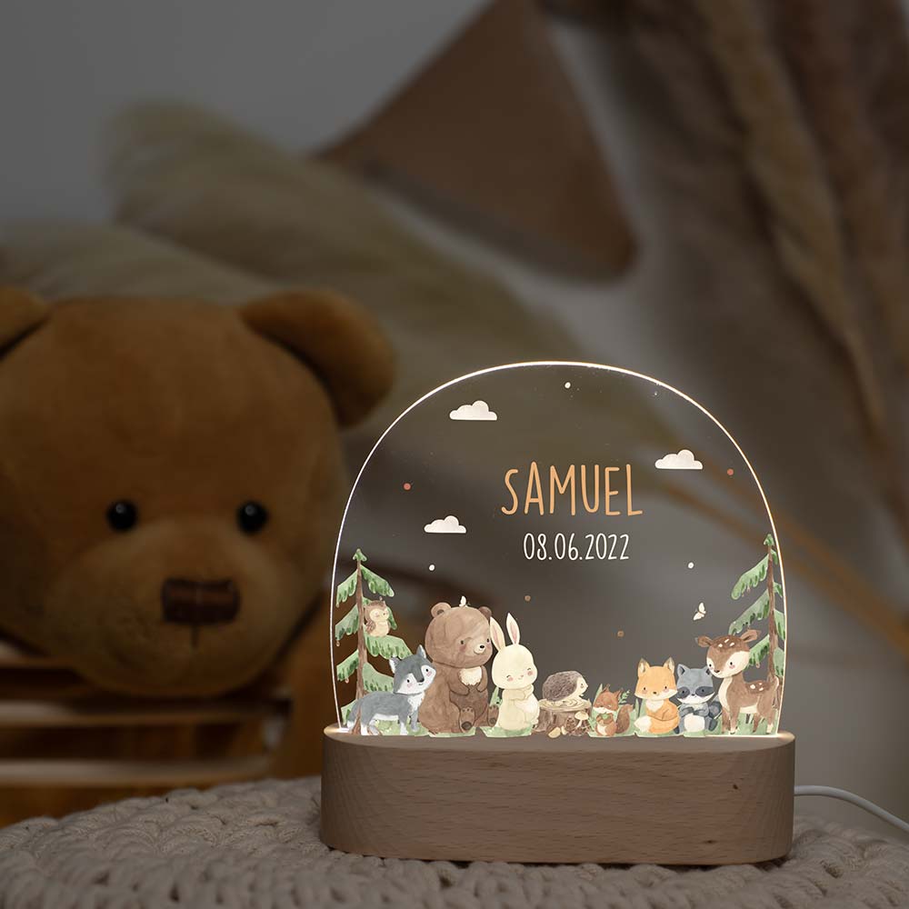 Personalisiertes Kindernachtlicht mit Waldtier-Motiv auf Holzsockel. Rundes, beleuchtetes Display zeigt niedliche Waldtiere unter dem Namen "SAMUEL" und Geburtsdatum. Im Hintergrund ein Teddybär. Perfektes Geschenk für Babys und Kinder.