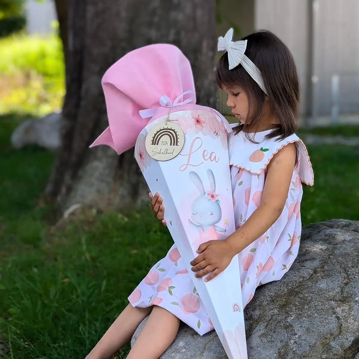 Mädchen mit personalisierter Schultüte zur Einschulung - Kind im hellen Sommerkleid mit Haarschleife betrachtet liebevoll ihre rosa-weiße Zuckertüte mit Häschen-Motiv und dem Namen 'Lea' im Garten
